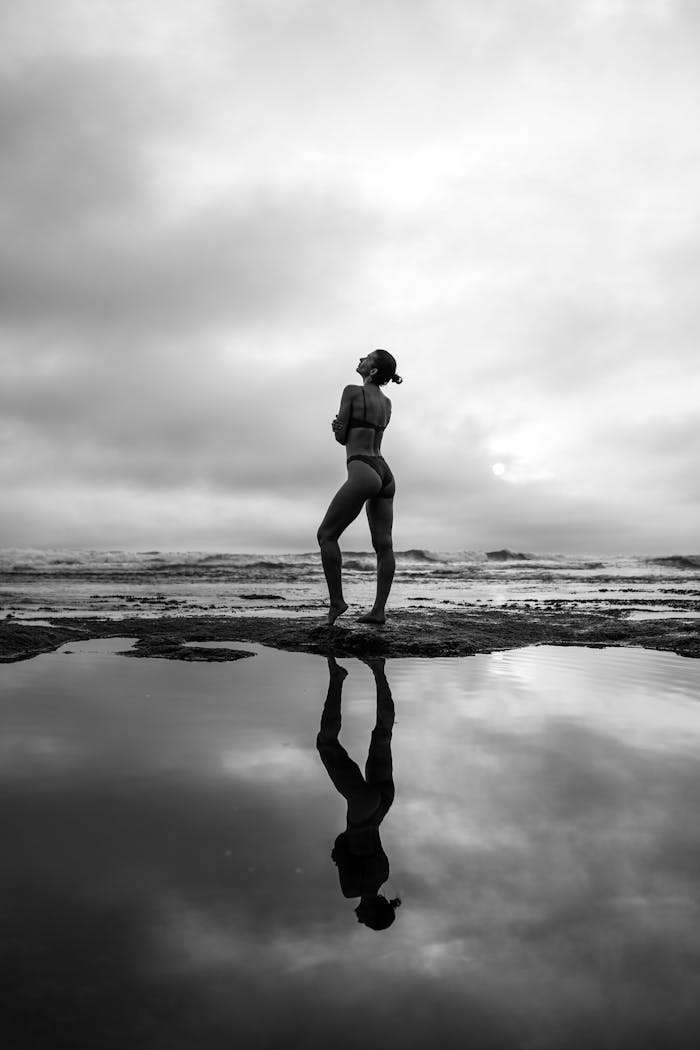 Breathtaking black and white shot of a woman by a reflective water pool under a cloudy sky.